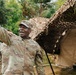 A U.S. Army soldier with the name tape 'SALAMI' and a Command Sergeant Major rank insignia is gesturing with his right arm raised, appearing to be speaking or giving instructions in a field environment. He is surrounded by other soldiers in OCP (Operational Camouflage Pattern) uniforms near a military field tent covered with camouflage netting. The setting appears to be an outdoor military training or operational area with trees in the background.