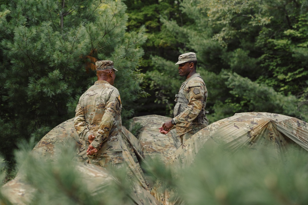 Two U.S. Army soldiers in OCP (Operational Camouflage Pattern) uniforms are working with what appears to be a large camouflage tarp or tent material in a wooded outdoor setting. The soldier on the right is facing forward and appears to be speaking or giving instruction, while the soldier on the left has his back to the camera. They appear to be engaged in field training or camp setup activities.
