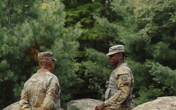 Two U.S. Army soldiers in OCP (Operational Camouflage Pattern) uniforms are working with what appears to be a large camouflage tarp or tent material in a wooded outdoor setting. The soldier on the right is facing forward and appears to be speaking or giving instruction, while the soldier on the left has his back to the camera. They appear to be engaged in field training or camp setup activities.