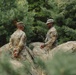 Two U.S. Army soldiers in OCP (Operational Camouflage Pattern) uniforms are working with what appears to be a large camouflage tarp or tent material in a wooded outdoor setting. The soldier on the right is facing forward and appears to be speaking or giving instruction, while the soldier on the left has his back to the camera. They appear to be engaged in field training or camp setup activities.