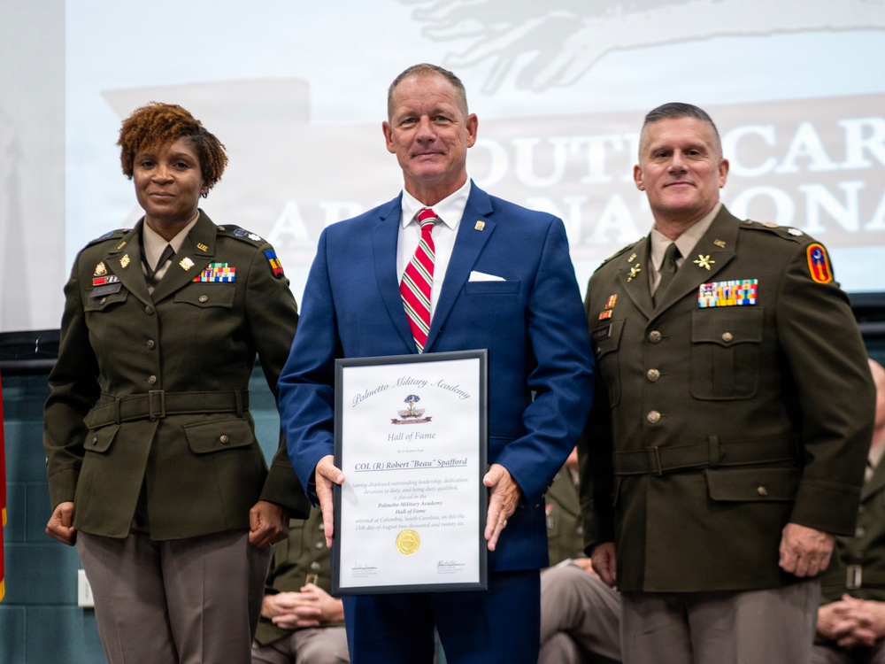 A formal military ceremony at Palmetto Military Academy where a civilian man in a blue suit is being inducted into the Hall of Fame. He is flanked by two military officers in Army dress uniforms and is holding a framed Hall of Fame certificate. The certificate is made out to 'COL (R) Robert "Beau" Spafford' and references Columbia, South Carolina.