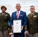 A formal military ceremony at Palmetto Military Academy where a civilian man in a blue suit is being inducted into the Hall of Fame. He is flanked by two military officers in Army dress uniforms and is holding a framed Hall of Fame certificate. The certificate is made out to 'COL (R) Robert "Beau" Spafford' and references Columbia, South Carolina.