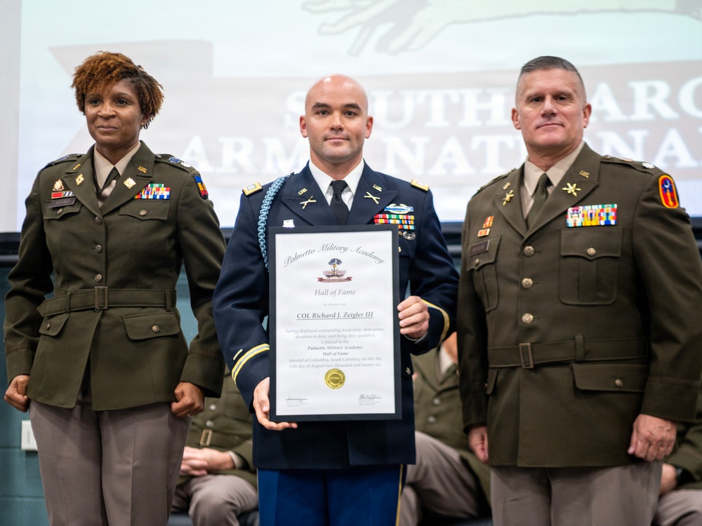 Three military personnel are posing for a formal photograph at what appears to be a Palmetto Military Academy Hall of Fame induction ceremony. The central figure, identified on the certificate as COL Richard J. Zeigler III, is holding a framed Hall of Fame certificate. The event appears to be held in South Carolina, as indicated by the 'South Carolina Army National Guard' banner visible in the background.