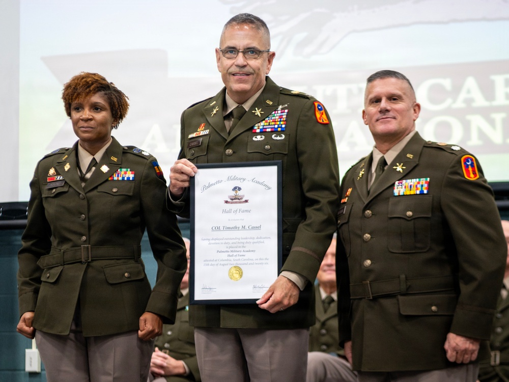 Three military personnel in U.S. Army dress uniforms pose together at what appears to be a formal ceremony. The central figure, identified on the certificate as COL Timothy M. Cassel, is holding a Palmetto Military Academy Hall of Fame induction certificate. The event appears to be taking place at a military academy in Columbia, South Carolina.