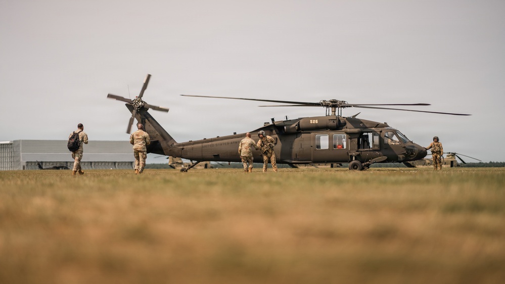 A United States Army UH-60 Black Hawk helicopter (tail number 526) is grounded on a grass field while several soldiers in camouflage uniforms approach or interact with the aircraft. The scene appears to depict a military loading or briefing operation, likely during a training exercise or operational deployment. A second helicopter is faintly visible in the background to the right.
