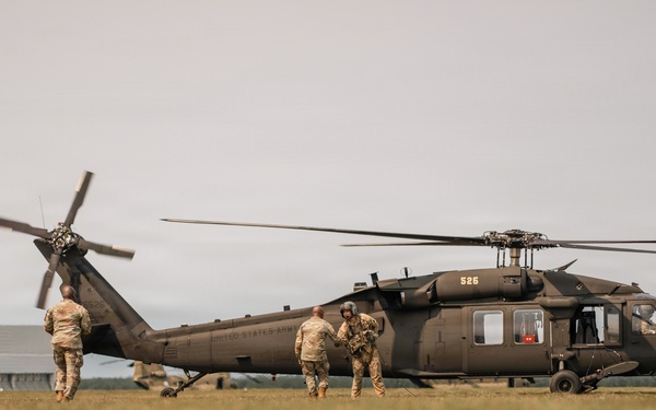 A United States Army UH-60 Black Hawk helicopter (tail number 526) is grounded on a grass field while several soldiers in camouflage uniforms approach or interact with the aircraft. The scene appears to depict a military loading or briefing operation, likely during a training exercise or operational deployment. A second helicopter is faintly visible in the background to the right.