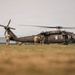 A United States Army UH-60 Black Hawk helicopter (tail number 526) is grounded on a grass field while several soldiers in camouflage uniforms approach or interact with the aircraft. The scene appears to depict a military loading or briefing operation, likely during a training exercise or operational deployment. A second helicopter is faintly visible in the background to the right.