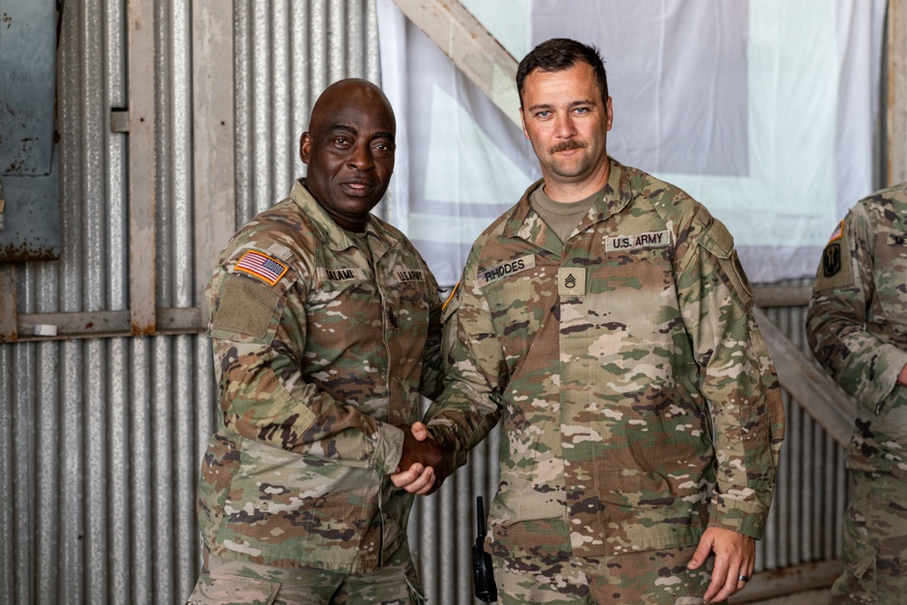 Two U.S. Army soldiers in Operational Camouflage Pattern (OCP) uniforms are shaking hands and posing for a photo inside what appears to be a metal-walled military facility or hangar. The soldier on the right has a name tape reading 'RHODES' and displays Staff Sergeant rank insignia, while the soldier on the left has a name tape that appears to read 'ALAMI' or similar. A third soldier is partially visible on the right edge of the frame.