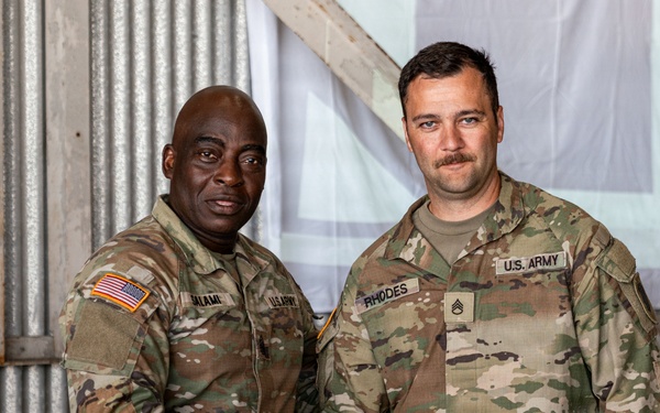 Two U.S. Army soldiers in Operational Camouflage Pattern (OCP) uniforms are shaking hands and posing for a photo inside what appears to be a metal-walled military facility or hangar. The soldier on the right has a name tape reading 'RHODES' and displays Staff Sergeant rank insignia, while the soldier on the left has a name tape that appears to read 'ALAMI' or similar. A third soldier is partially visible on the right edge of the frame.