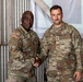 Two U.S. Army soldiers in Operational Camouflage Pattern (OCP) uniforms are shaking hands and posing for a photo inside what appears to be a metal-walled military facility or hangar. The soldier on the right has a name tape reading 'RHODES' and displays Staff Sergeant rank insignia, while the soldier on the left has a name tape that appears to read 'ALAMI' or similar. A third soldier is partially visible on the right edge of the frame.