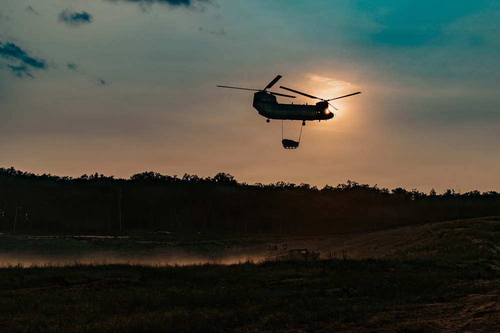 A tandem-rotor helicopter, consistent with a CH-47 Chinook, is silhouetted against a sunset sky while performing a sling-load operation, carrying a suspended cargo load beneath its fuselage. The scene appears to be a military training or operational environment, with open terrain, dust disturbance on the ground, and at least one ground vehicle visible below. The dramatic backlighting from the setting sun creates a strong silhouette effect across the entire scene.