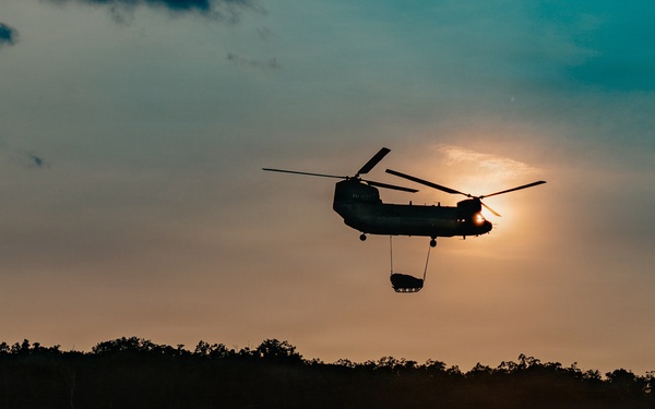 A tandem-rotor helicopter, consistent with a CH-47 Chinook, is silhouetted against a sunset sky while performing a sling-load operation, carrying a suspended cargo load beneath its fuselage. The scene appears to be a military training or operational environment, with open terrain, dust disturbance on the ground, and at least one ground vehicle visible below. The dramatic backlighting from the setting sun creates a strong silhouette effect across the entire scene.