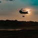 A tandem-rotor helicopter, consistent with a CH-47 Chinook, is silhouetted against a sunset sky while performing a sling-load operation, carrying a suspended cargo load beneath its fuselage. The scene appears to be a military training or operational environment, with open terrain, dust disturbance on the ground, and at least one ground vehicle visible below. The dramatic backlighting from the setting sun creates a strong silhouette effect across the entire scene.