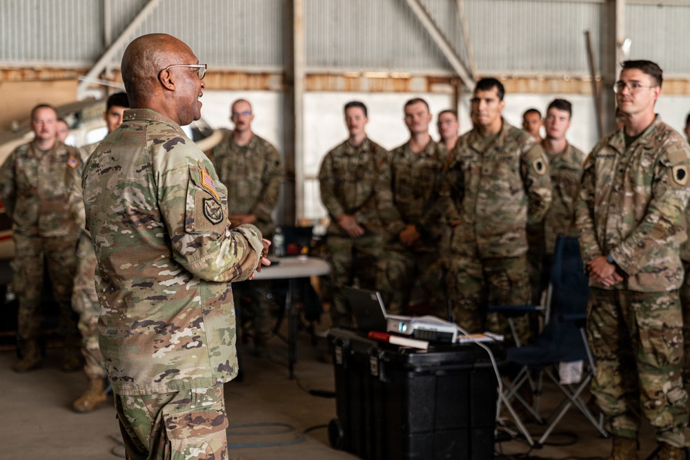 A senior military officer in OCP (Operational Camouflage Pattern) uniform is addressing a group of assembled soldiers/airmen inside what appears to be a military hangar. The speaker stands with his back to the camera, facing a formation of approximately 15-20 personnel who are listening attentively. A laptop and equipment case are visible in the foreground, suggesting a briefing or training session.
