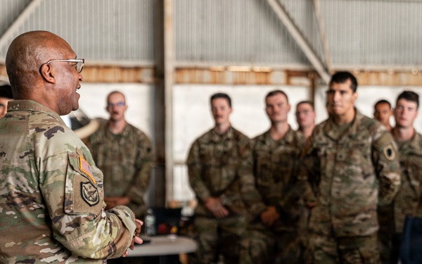 A senior military officer in OCP (Operational Camouflage Pattern) uniform is addressing a group of assembled soldiers/airmen inside what appears to be a military hangar. The speaker stands with his back to the camera, facing a formation of approximately 15-20 personnel who are listening attentively. A laptop and equipment case are visible in the foreground, suggesting a briefing or training session.
