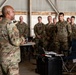 A senior military officer in OCP (Operational Camouflage Pattern) uniform is addressing a group of assembled soldiers/airmen inside what appears to be a military hangar. The speaker stands with his back to the camera, facing a formation of approximately 15-20 personnel who are listening attentively. A laptop and equipment case are visible in the foreground, suggesting a briefing or training session.