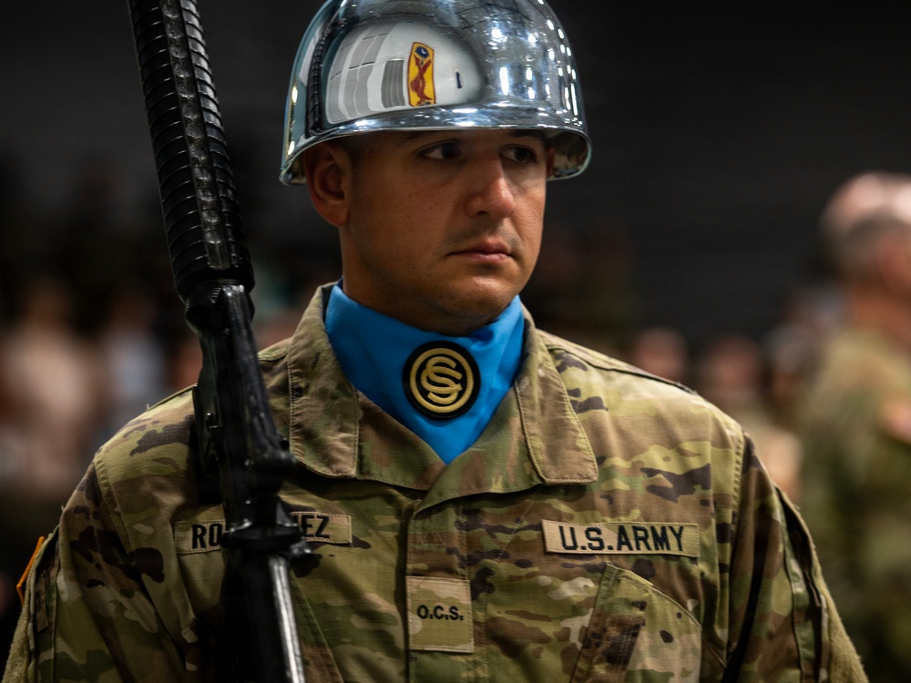 A U.S. Army soldier in ceremonial dress stands at attention during what appears to be an Officer Candidate School (OCS) ceremony. The soldier wears a distinctive chrome/silver helmet, blue ascot with OCS medallion, and OCP (Operational Camouflage Pattern) uniform while holding a rifle with bayonet. The name tape reads 'RODRIGUEZ' and unit tape reads 'O.C.S.'