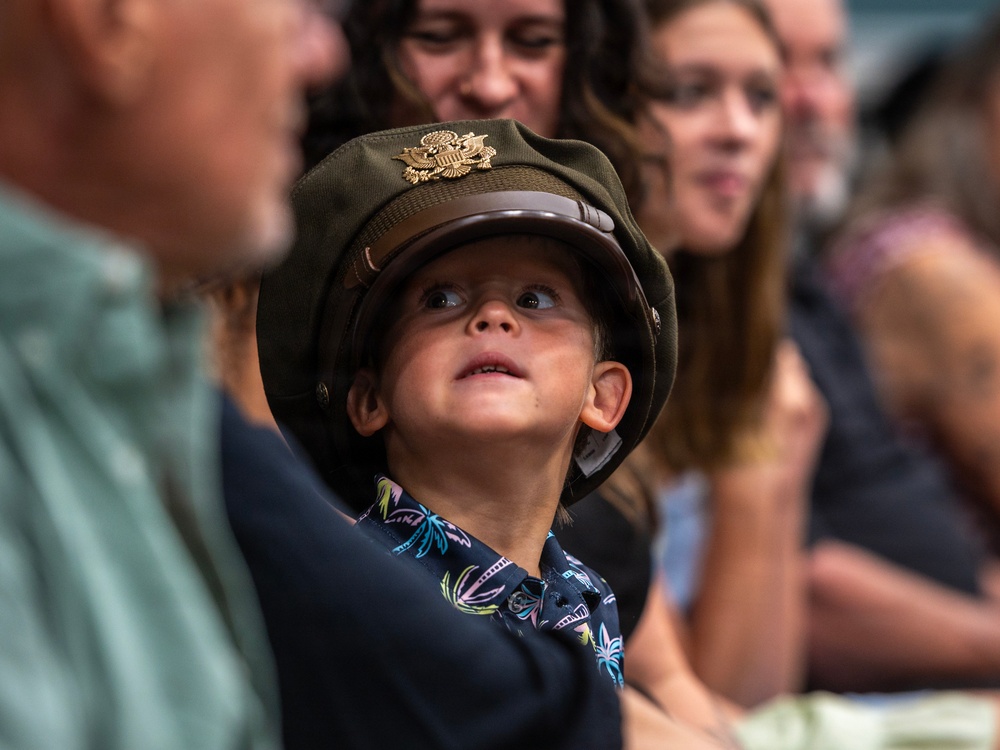 A young child wearing an oversized olive-green U.S. military officer's cap with a gold eagle insignia looks upward with wide, curious eyes. The child is dressed in a colorful tropical-print shirt and is surrounded by adults in what appears to be an indoor gathering or ceremony. The image captures a candid, endearing moment likely at a military-related event.
