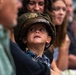 A young child wearing an oversized olive-green U.S. military officer's cap with a gold eagle insignia looks upward with wide, curious eyes. The child is dressed in a colorful tropical-print shirt and is surrounded by adults in what appears to be an indoor gathering or ceremony. The image captures a candid, endearing moment likely at a military-related event.