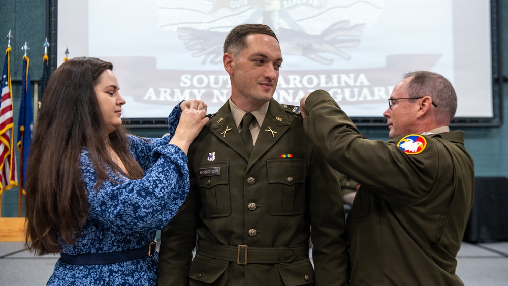 A military promotion ceremony for a South Carolina Army National Guard soldier named Bryngelson, during which new rank insignia are being pinned onto his shoulders. A woman in a blue floral dress pins one shoulder while an older male officer in Army greens pins the other. The event takes place in a formal indoor setting with a projected backdrop displaying the South Carolina Army National Guard emblem.