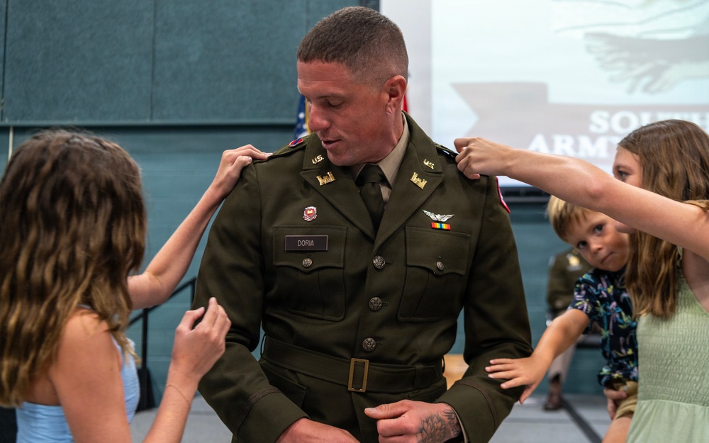 A U.S. Army officer named Doria is having rank insignia pinned onto his shoulders by children during what appears to be a military promotion ceremony. The setting includes a backdrop displaying 'SOUTH ARMY' (partially visible) and an American flag. This is a traditional 'pinning ceremony' where family members, typically children or spouses, attach the new rank insignia to the promoted officer's uniform.