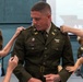 A U.S. Army officer named Doria is having rank insignia pinned onto his shoulders by children during what appears to be a military promotion ceremony. The setting includes a backdrop displaying 'SOUTH ARMY' (partially visible) and an American flag. This is a traditional 'pinning ceremony' where family members, typically children or spouses, attach the new rank insignia to the promoted officer's uniform.