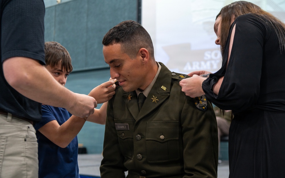 A U.S. Army officer named Suarez is having rank insignia pinned onto his uniform collar during what appears to be a military promotion ceremony. Family members, including a young boy and a woman in a black dress, are participating in the traditional pinning ritual. The ceremony appears to take place in an indoor venue with Army-related signage visible in the background.