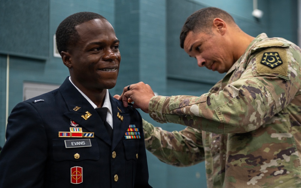 A military promotion or award ceremony is taking place, with one soldier in Army Service Uniform (dress blues) named Evans receiving a pin or insignia on his collar from another soldier in Army Combat Uniform (OCP/MultiCam). The soldier being pinned is smiling broadly, suggesting a positive milestone event. Both individuals appear to be U.S. Army personnel based on their uniforms and insignia.
