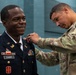 A military promotion or award ceremony is taking place, with one soldier in Army Service Uniform (dress blues) named Evans receiving a pin or insignia on his collar from another soldier in Army Combat Uniform (OCP/MultiCam). The soldier being pinned is smiling broadly, suggesting a positive milestone event. Both individuals appear to be U.S. Army personnel based on their uniforms and insignia.