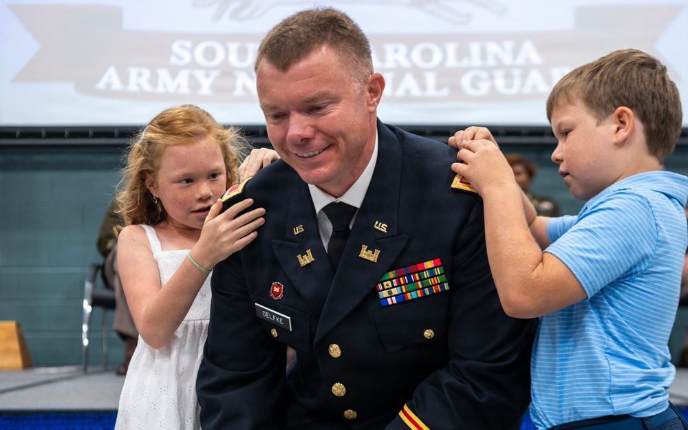 A U.S. Army officer named Oelfke, wearing dress blues, is having rank insignia pinned onto his shoulders by two children during what appears to be a promotion ceremony for the South Carolina Army National Guard. The officer is smiling broadly as a young red-haired girl pins his left shoulder and a boy in a blue striped shirt pins his right. The setting is an official military ceremony with a South Carolina Army National Guard banner visible in the background.