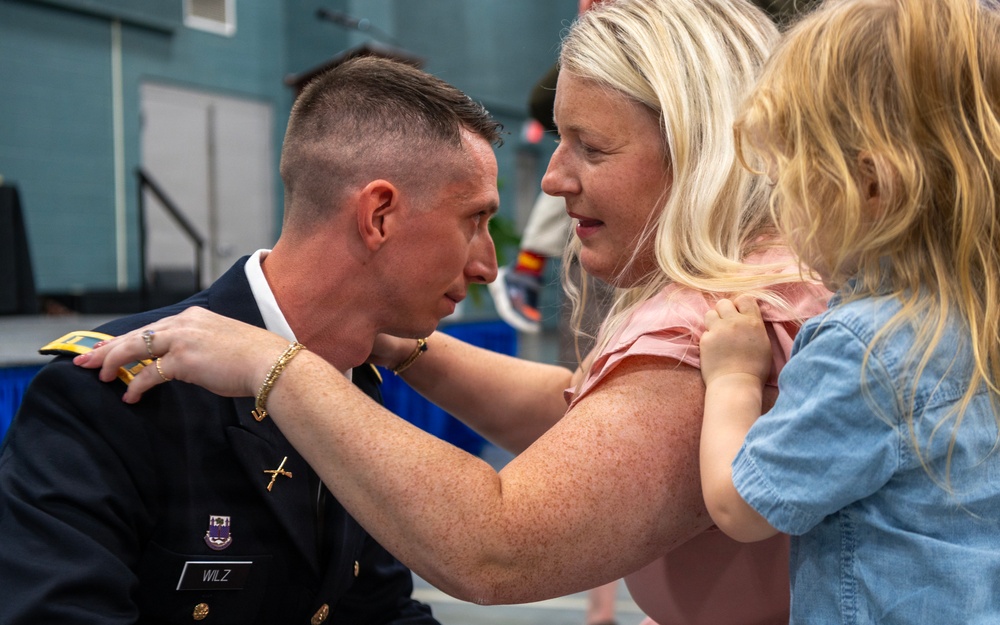 A U.S. Army soldier named WILZ, wearing dress blues, is leaning forward in what appears to be a promotion or pinning ceremony. A blonde woman, likely his spouse, is pinning a new insignia or rank onto his uniform while a young blonde child in a denim shirt looks on. The scene takes place in what appears to be a military event hall or gymnasium.