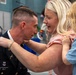 A U.S. Army soldier named WILZ, wearing dress blues, is leaning forward in what appears to be a promotion or pinning ceremony. A blonde woman, likely his spouse, is pinning a new insignia or rank onto his uniform while a young blonde child in a denim shirt looks on. The scene takes place in what appears to be a military event hall or gymnasium.