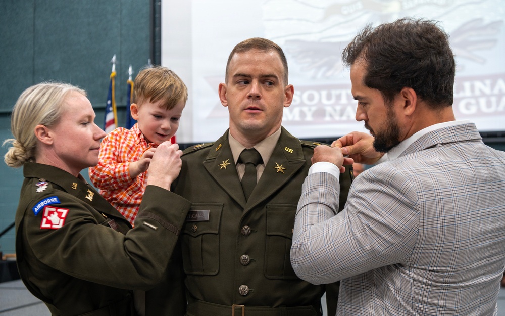 A military promotion ceremony is taking place, likely within the South Carolina Army National Guard based on the projection screen in the background. A soldier named Thomas is having new rank insignia pinned onto his uniform collar by two individuals — a female soldier in Army greens holding a young child, and a civilian man in a plaid blazer. The ceremony appears to be an intimate, formal occasion with family participation.