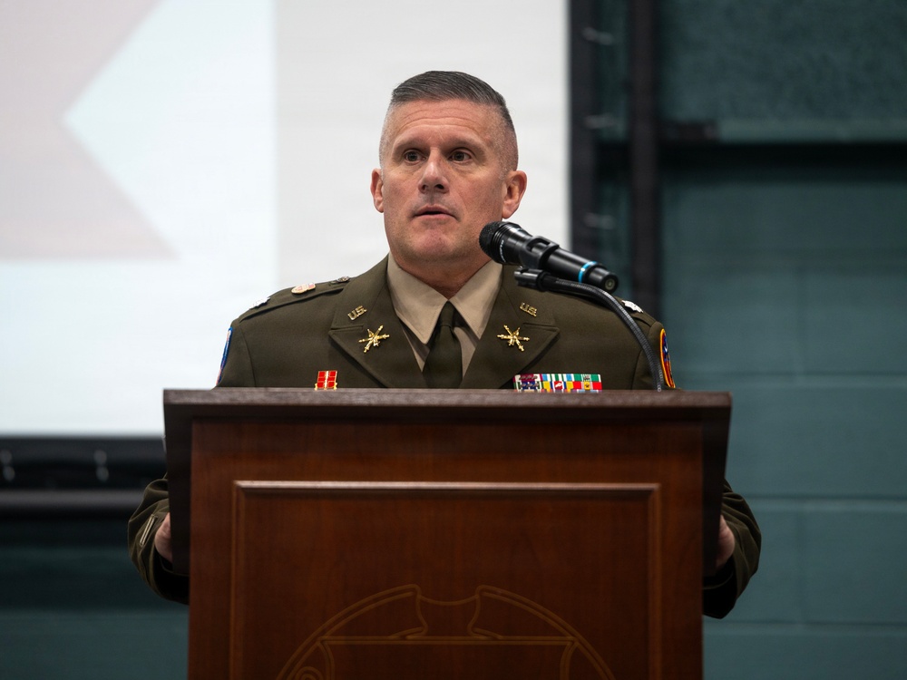 A U.S. Army officer in dress uniform is speaking at a wooden podium equipped with a microphone. The officer displays multiple service ribbons, medals, and branch insignia on their uniform, suggesting a senior rank. The setting appears to be an indoor military ceremony or formal briefing.