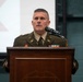 A U.S. Army officer in dress uniform is speaking at a wooden podium equipped with a microphone. The officer displays multiple service ribbons, medals, and branch insignia on their uniform, suggesting a senior rank. The setting appears to be an indoor military ceremony or formal briefing.
