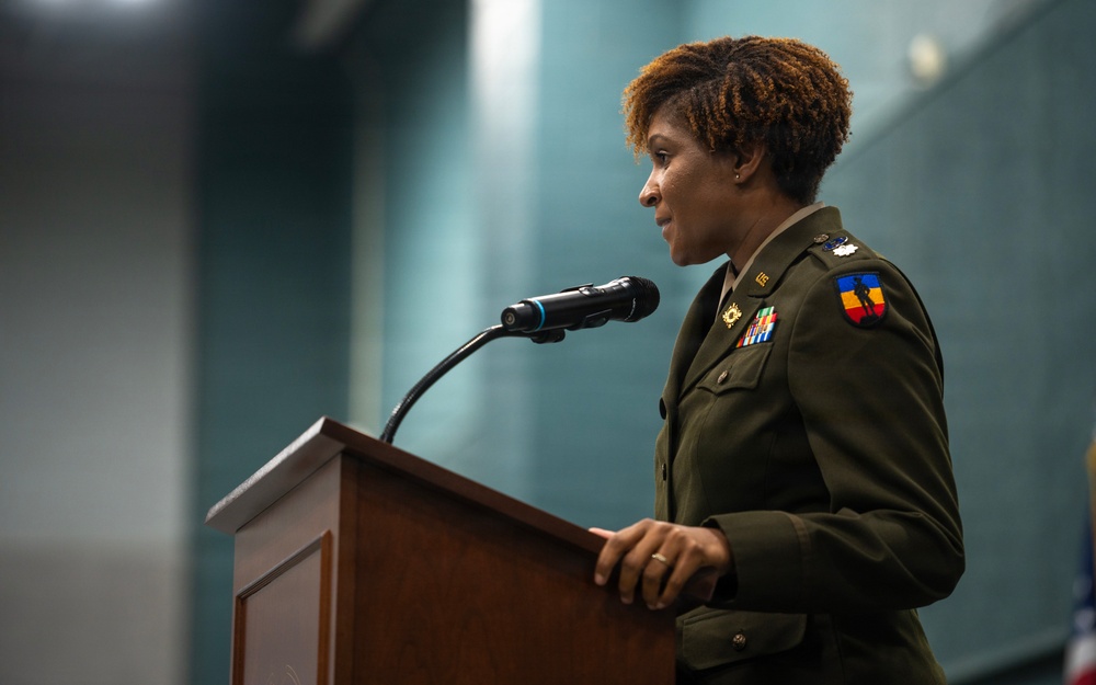 A female military officer in U.S. Army dress uniform is speaking at a wooden podium equipped with a microphone. She is wearing an olive green Army Service Uniform with visible rank insignia, unit patch, and service ribbons. The setting appears to be an indoor formal military ceremony or official event.