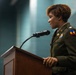 A female military officer in U.S. Army dress uniform is speaking at a wooden podium equipped with a microphone. She is wearing an olive green Army Service Uniform with visible rank insignia, unit patch, and service ribbons. The setting appears to be an indoor formal military ceremony or official event.
