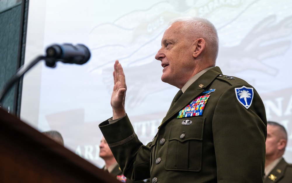 A senior U.S. Army officer in dress green uniform is raising his right hand, appearing to take an oath or be sworn in at a formal proceeding. He stands at a podium with a microphone, wearing numerous service ribbons, badges, and a South Carolina National Guard shoulder patch. Other uniformed personnel are visible in the background.