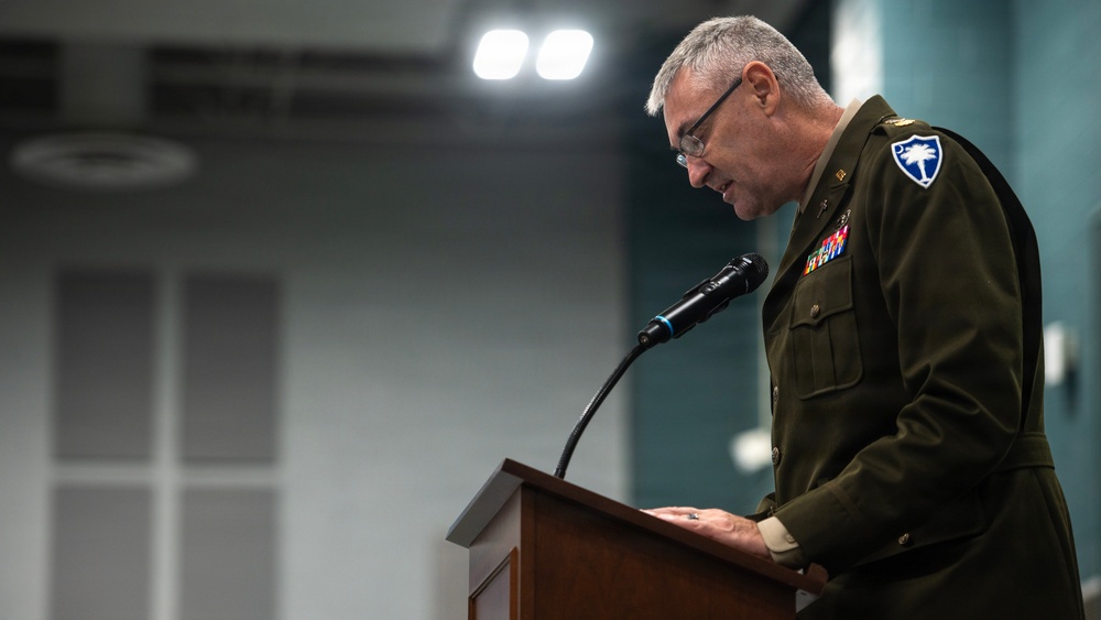 A senior military officer in dress uniform stands at a wooden podium speaking into a microphone, appearing to read from notes. He is wearing a dark olive/green Army dress uniform adorned with multiple service ribbons and medals, along with a shoulder patch visible on his left sleeve. The setting appears to be an official military briefing room or auditorium.