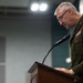 A senior military officer in dress uniform stands at a wooden podium speaking into a microphone, appearing to read from notes. He is wearing a dark olive/green Army dress uniform adorned with multiple service ribbons and medals, along with a shoulder patch visible on his left sleeve. The setting appears to be an official military briefing room or auditorium.