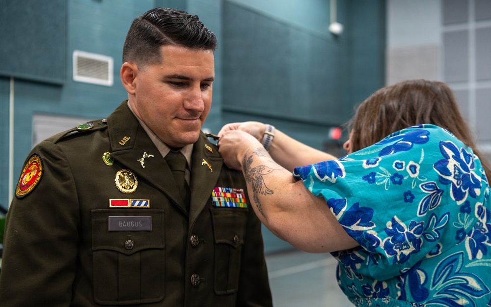 A military promotion or award ceremony is taking place, where a woman in a floral blue dress is pinning a new insignia or rank onto the uniform of a U.S. Army soldier identified by his name tag as 'BAUGUS'. The soldier is wearing an olive green Army Service Uniform adorned with multiple ribbons, badges, and patches including a U.S. Marine Corps patch. The soldier is smiling downward as the pinning occurs, suggesting this is a celebratory milestone event.