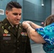 A military promotion or award ceremony is taking place, where a woman in a floral blue dress is pinning a new insignia or rank onto the uniform of a U.S. Army soldier identified by his name tag as 'BAUGUS'. The soldier is wearing an olive green Army Service Uniform adorned with multiple ribbons, badges, and patches including a U.S. Marine Corps patch. The soldier is smiling downward as the pinning occurs, suggesting this is a celebratory milestone event.