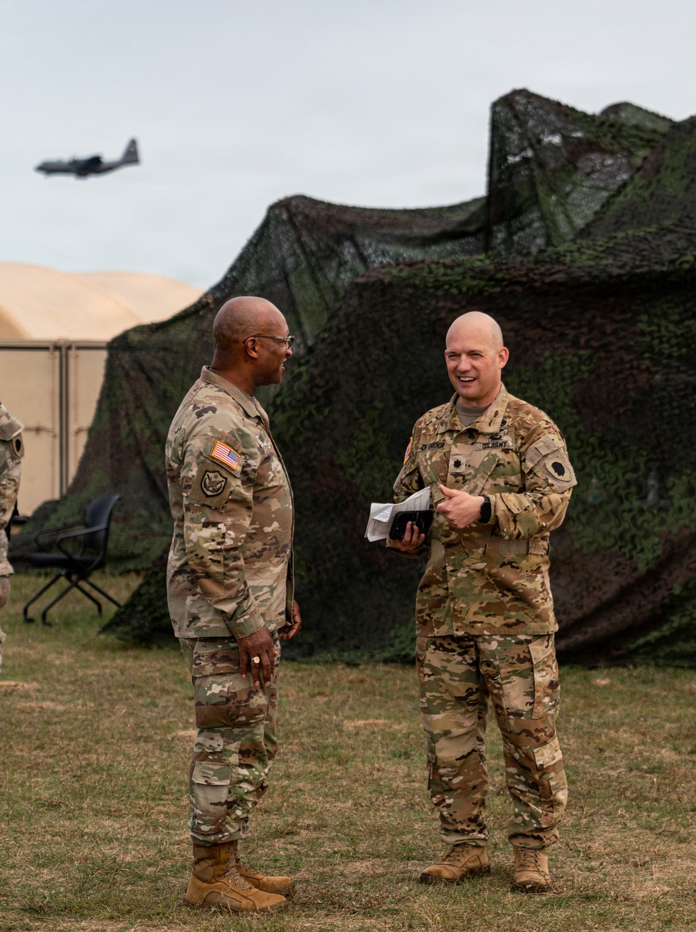 Two U.S. Army soldiers in Operational Camouflage Pattern (OCP) uniforms are engaged in conversation at what appears to be a field training exercise or forward operating base. A military aircraft, consistent with a C-130 Hercules, is visible in the background flying overhead. The setting includes camouflage netting, military tents, and a grassy field area.