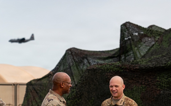 Two U.S. Army soldiers in Operational Camouflage Pattern (OCP) uniforms are engaged in conversation at what appears to be a field training exercise or forward operating base. A military aircraft, consistent with a C-130 Hercules, is visible in the background flying overhead. The setting includes camouflage netting, military tents, and a grassy field area.