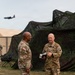 Two U.S. Army soldiers in Operational Camouflage Pattern (OCP) uniforms are engaged in conversation at what appears to be a field training exercise or forward operating base. A military aircraft, consistent with a C-130 Hercules, is visible in the background flying overhead. The setting includes camouflage netting, military tents, and a grassy field area.