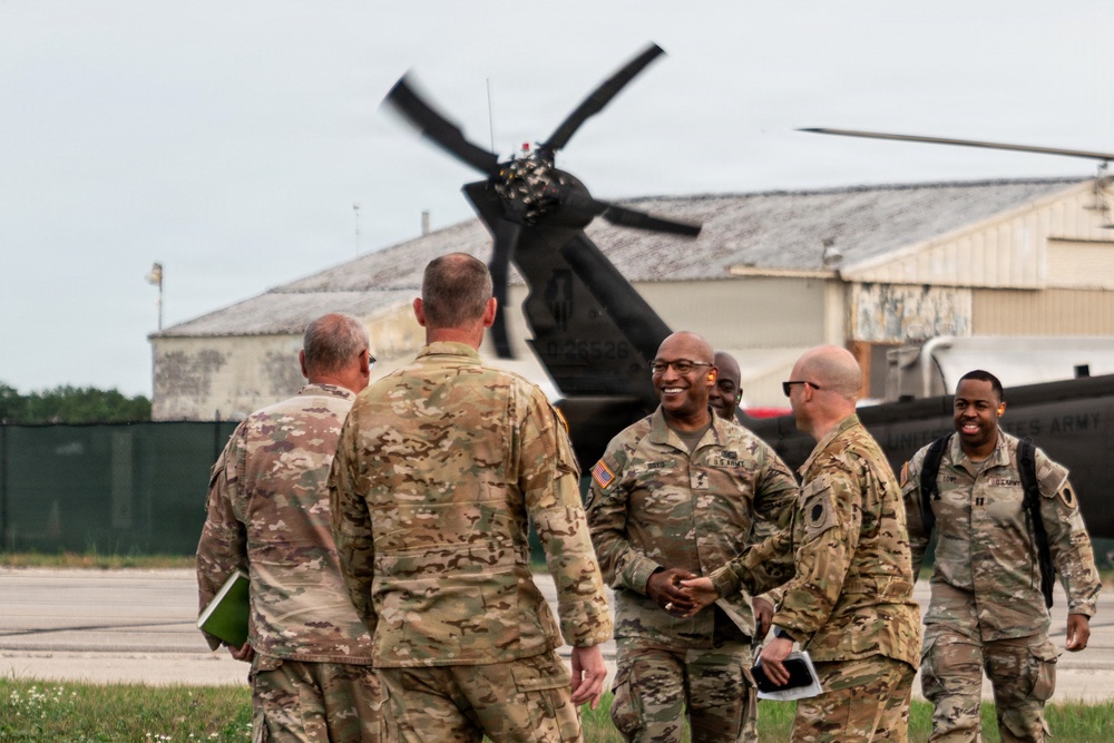 A group of U.S. Army soldiers in OCP (Operational Camouflage Pattern) uniforms are gathered on a military airfield tarmac, engaged in conversation with apparent camaraderie. A military helicopter with spinning rotors is visible in the background near what appears to be a hangar facility. The scene suggests an arrival, departure, or greeting ceremony at a military aviation installation.