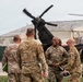 A group of U.S. Army soldiers in OCP (Operational Camouflage Pattern) uniforms are gathered on a military airfield tarmac, engaged in conversation with apparent camaraderie. A military helicopter with spinning rotors is visible in the background near what appears to be a hangar facility. The scene suggests an arrival, departure, or greeting ceremony at a military aviation installation.