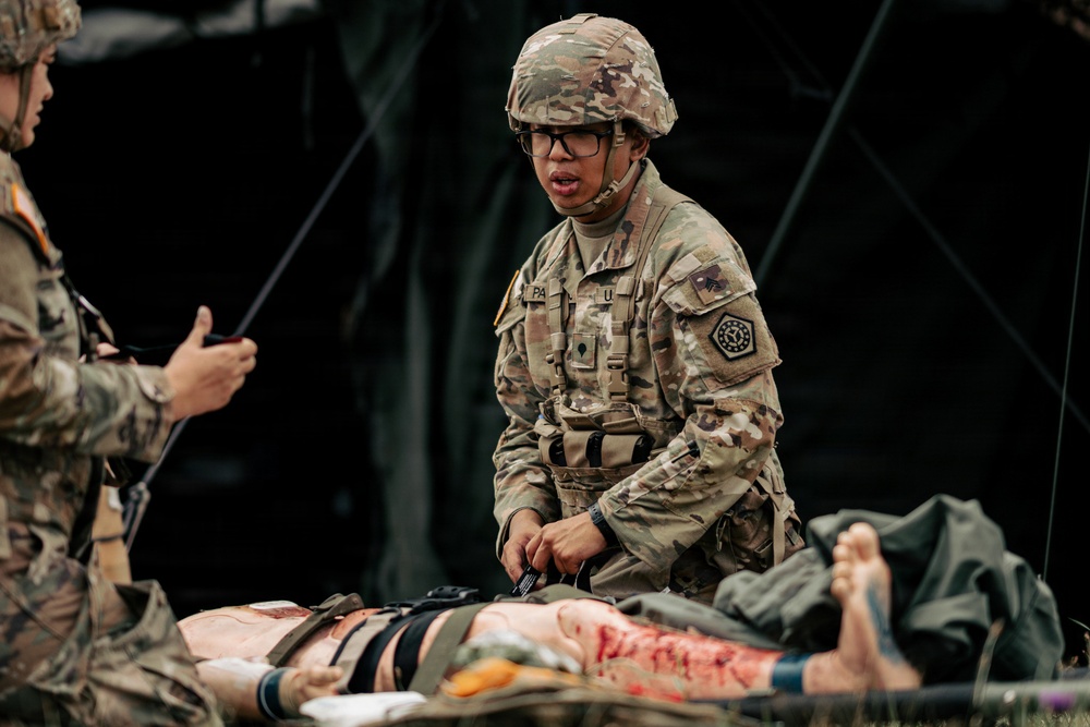 A U.S. Army soldier in full combat uniform and helmet is performing tactical combat casualty care (TCCC) on a simulated casualty during what appears to be a field medical training exercise. The soldier, wearing glasses and a name tape partially reading 'PA', is kneeling beside a moulage-equipped training mannequin or simulated casualty with realistic-looking wounds. A second uniformed soldier is visible on the left, and a military field tent is visible in the background.