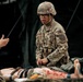 A U.S. Army soldier in full combat uniform and helmet is performing tactical combat casualty care (TCCC) on a simulated casualty during what appears to be a field medical training exercise. The soldier, wearing glasses and a name tape partially reading 'PA', is kneeling beside a moulage-equipped training mannequin or simulated casualty with realistic-looking wounds. A second uniformed soldier is visible on the left, and a military field tent is visible in the background.