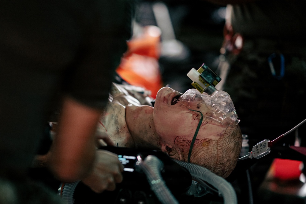 The image depicts what appears to be a medical training mannequin or simulation dummy with realistic wound makeup, surrounded by medical equipment and tubing. The figure has simulated blood and injuries on the head and neck area, consistent with a trauma training scenario. Multiple medical devices, tubes, and personnel hands are visible around the subject.